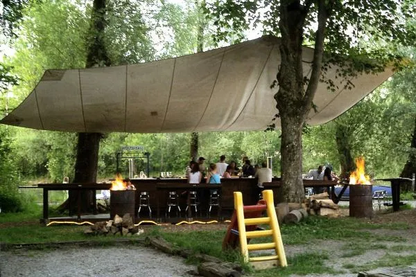 Bar de guinguette en plein air sous les arbres au bord de l'eau, tables et tabourets, lumière de fin d'après-midi